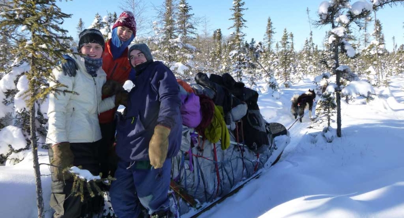 Three people smile while standing near a packed dog sled in a snowy wooded area. There are sled dogs attached to the sled. 